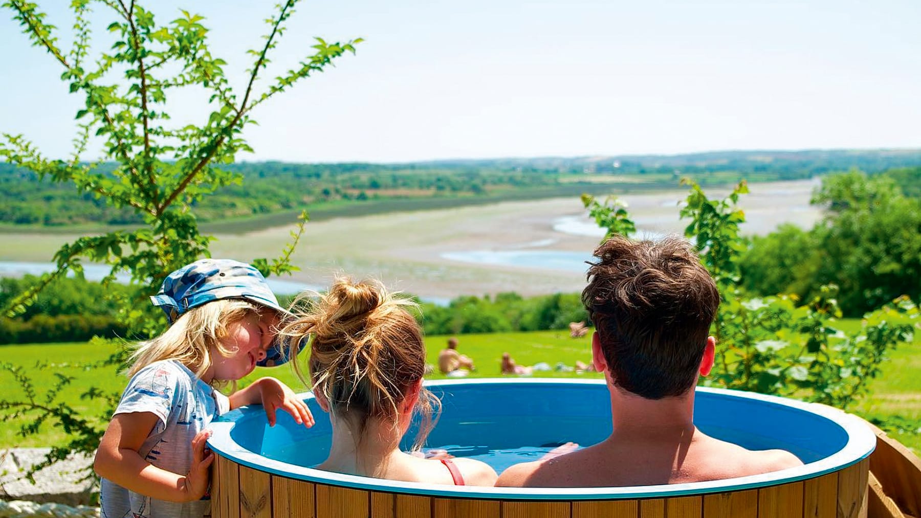 Photograph shows a couple in a hot tub overlooking a beach with a small child beside the hot tub