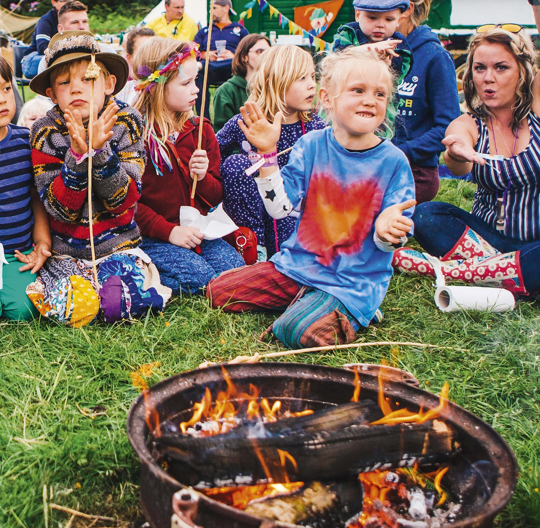Photograph shows children at a festival kneeling in front of a fire bowl with marshmallows on sticks