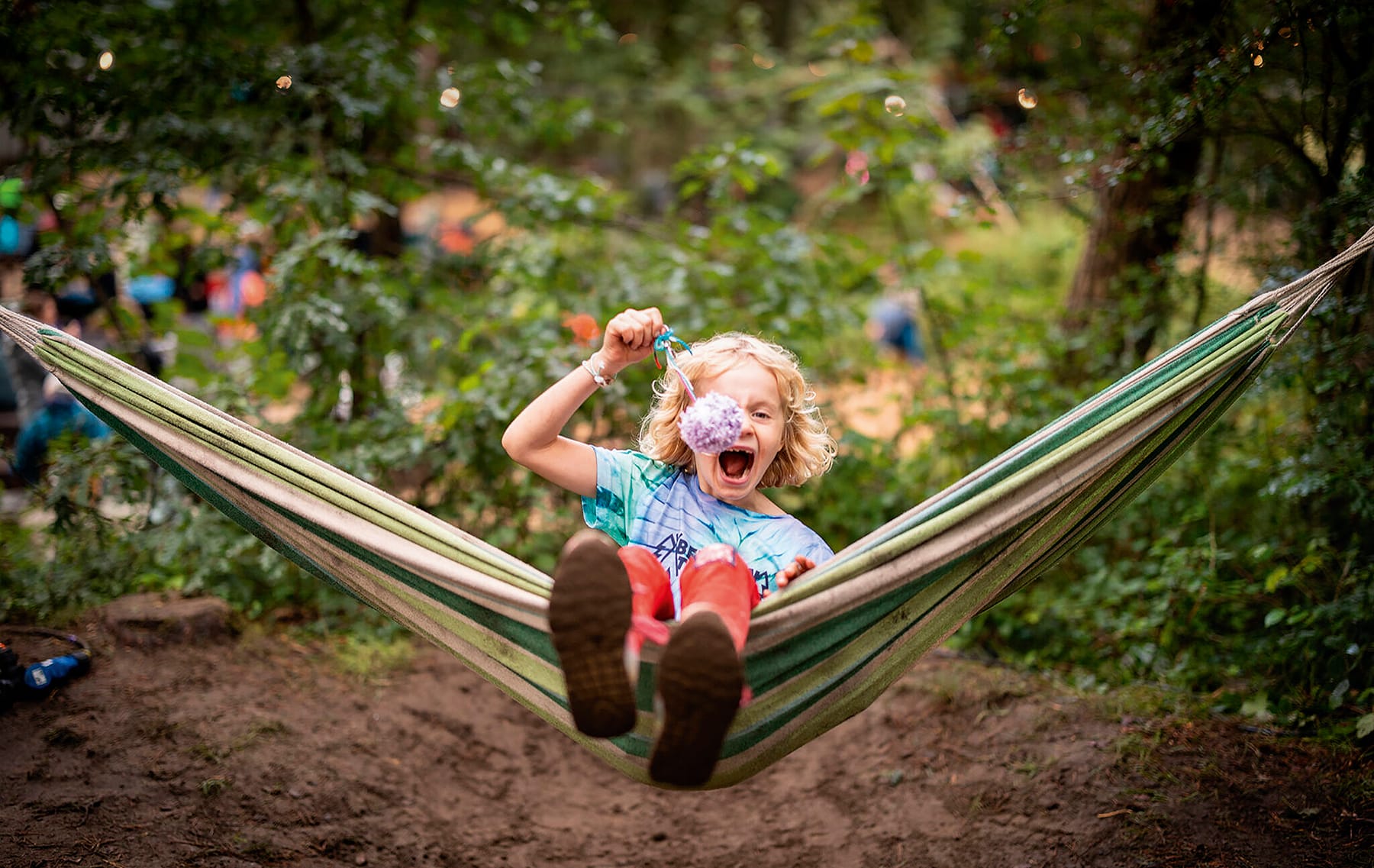 Photograph shows a young child at a festival, in a hammock holding a purple pompom
