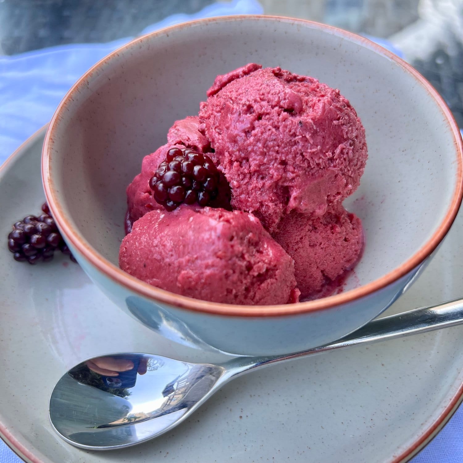 Photograph of a bowl of bright pink blackberry ice-cream served in a ceramic bowl with a spoon alongside