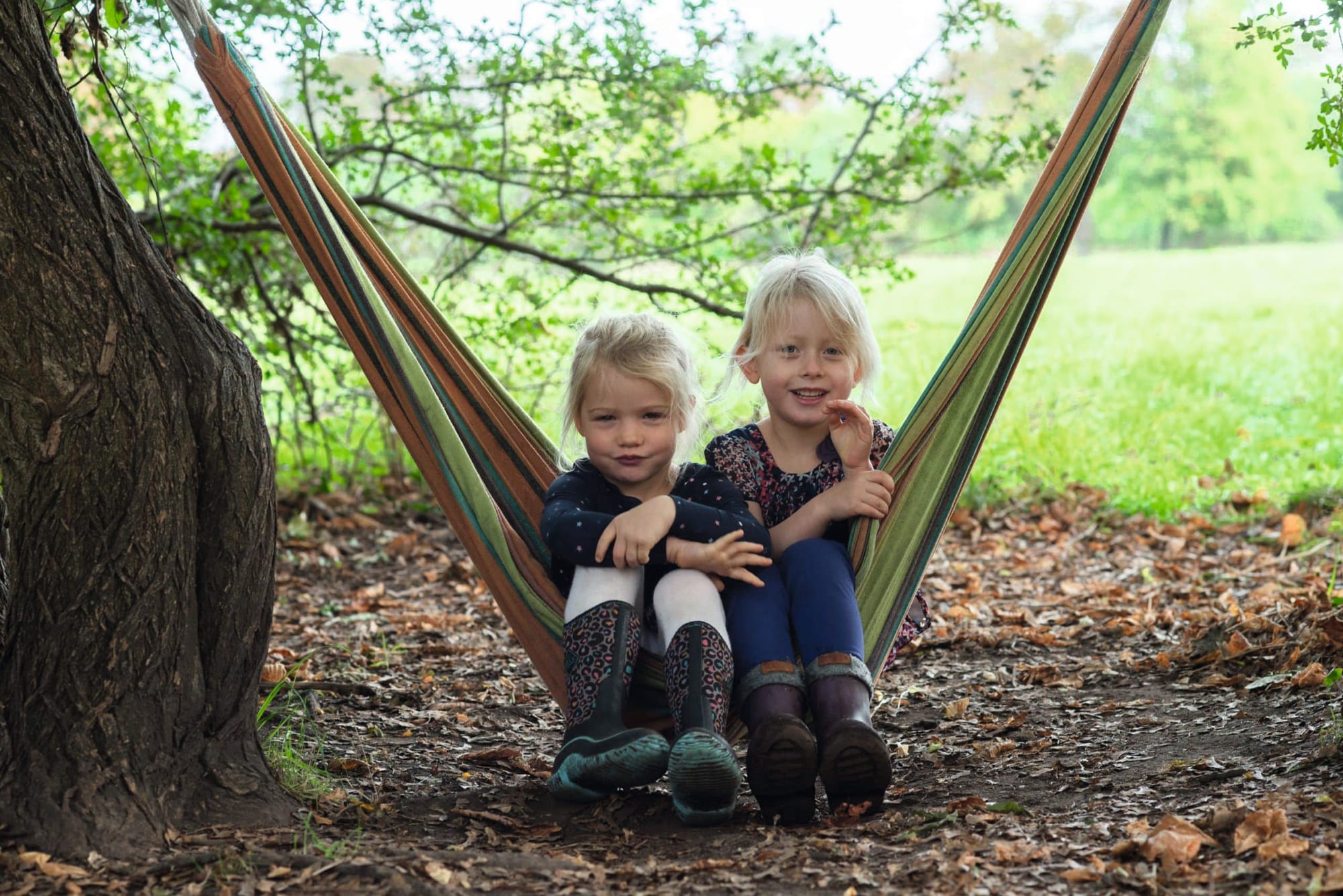 Photograph of two children sitting in a hammock strung from a large tree in the park