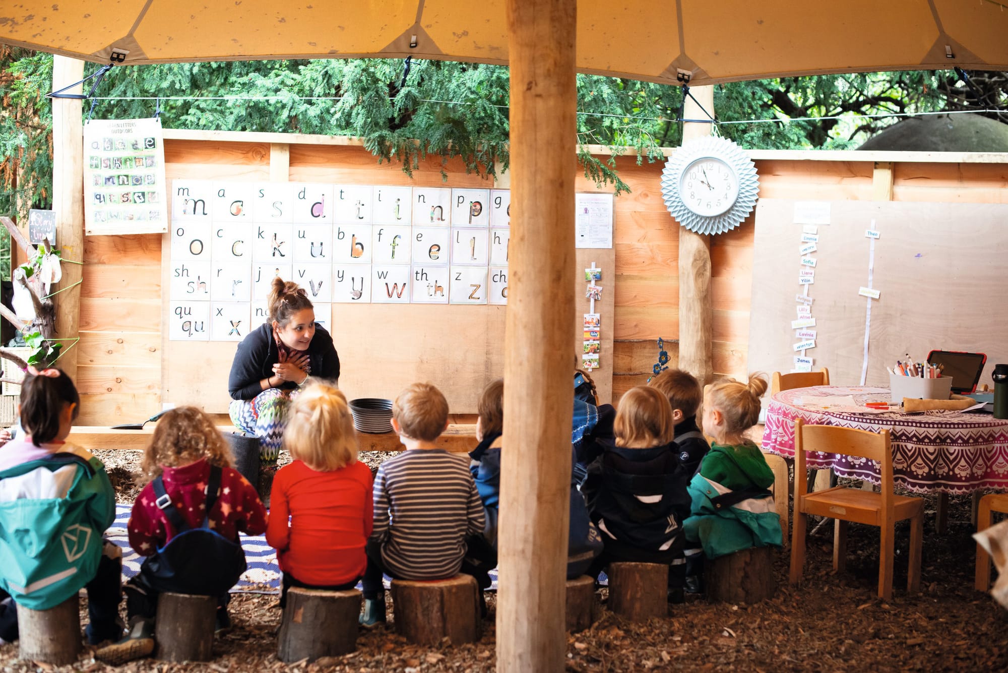 Photograph of a wooden outdoor classroom and children sitting on log seats listening to a teacher