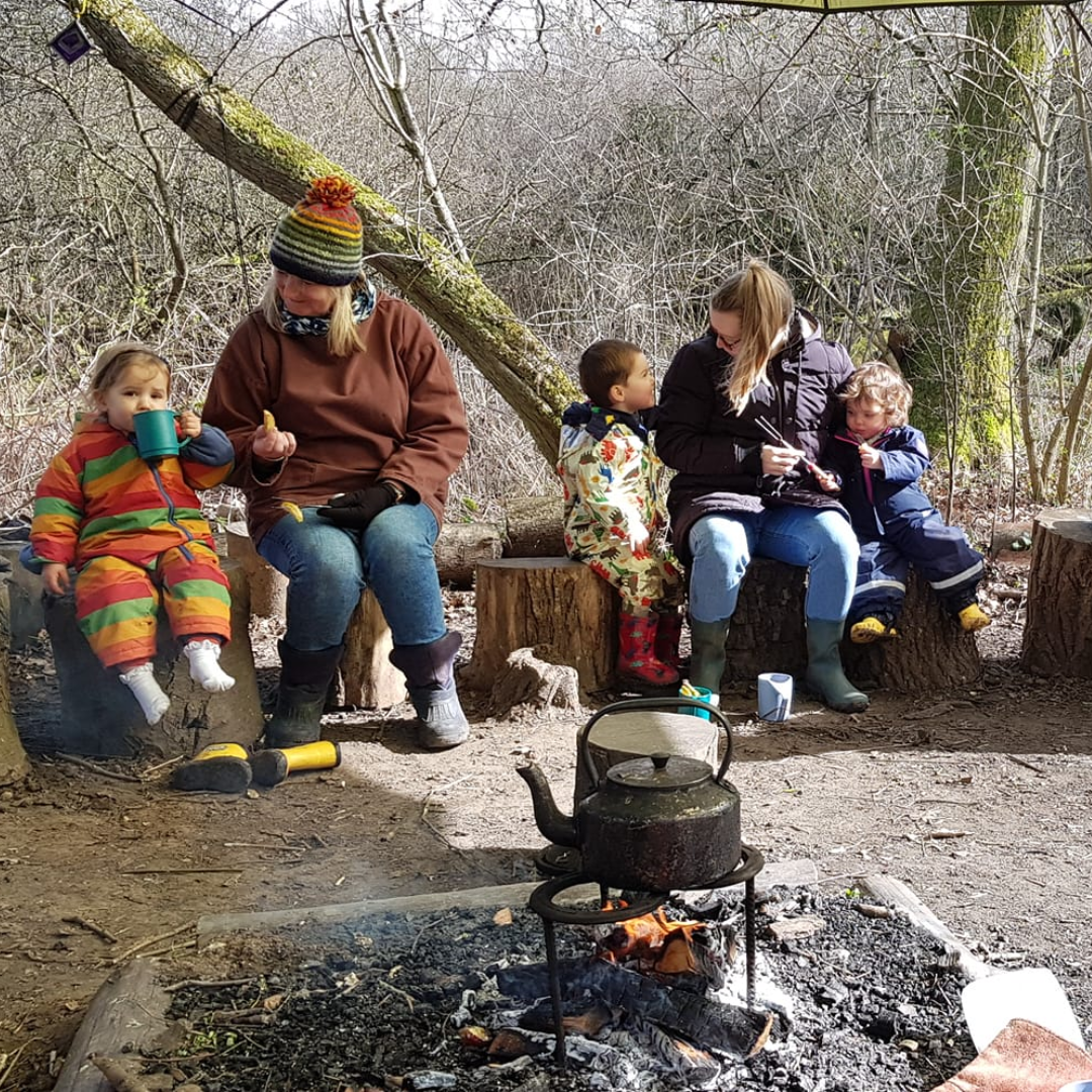 Photograph shows families sitting around a fire in the woods at Forest School with a kettle on a trivet in the centre