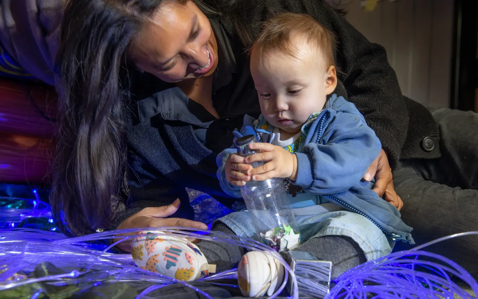 A baby sits with their mother. The baby exploring a bottle of paper stars while she looks on adoringly.