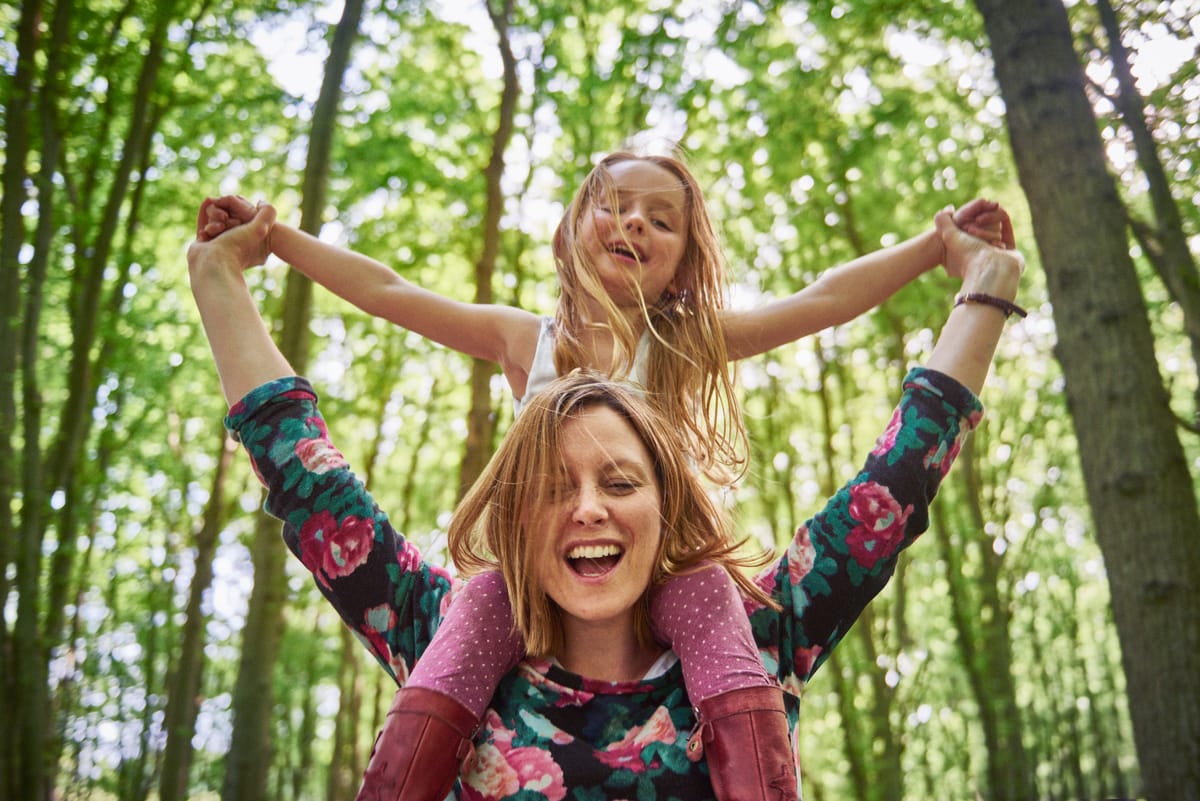 Photograph of a mother in the woods, with her daughter on her shoulders, arms raised and looking happy