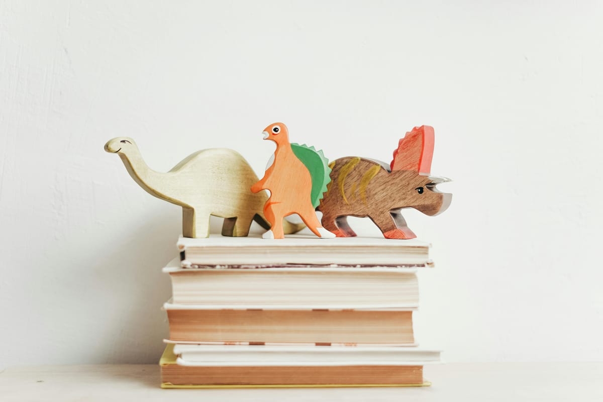 Photograph of a pile of books, spines in, with wooden dinosaurs balanced on top