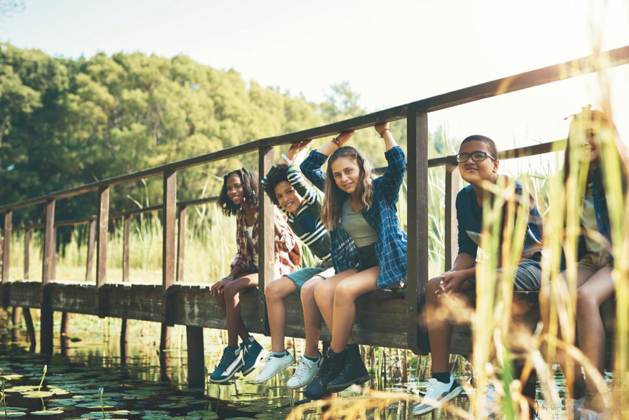Row of young people sitting on a wooden bridge over a lake in the sunshine