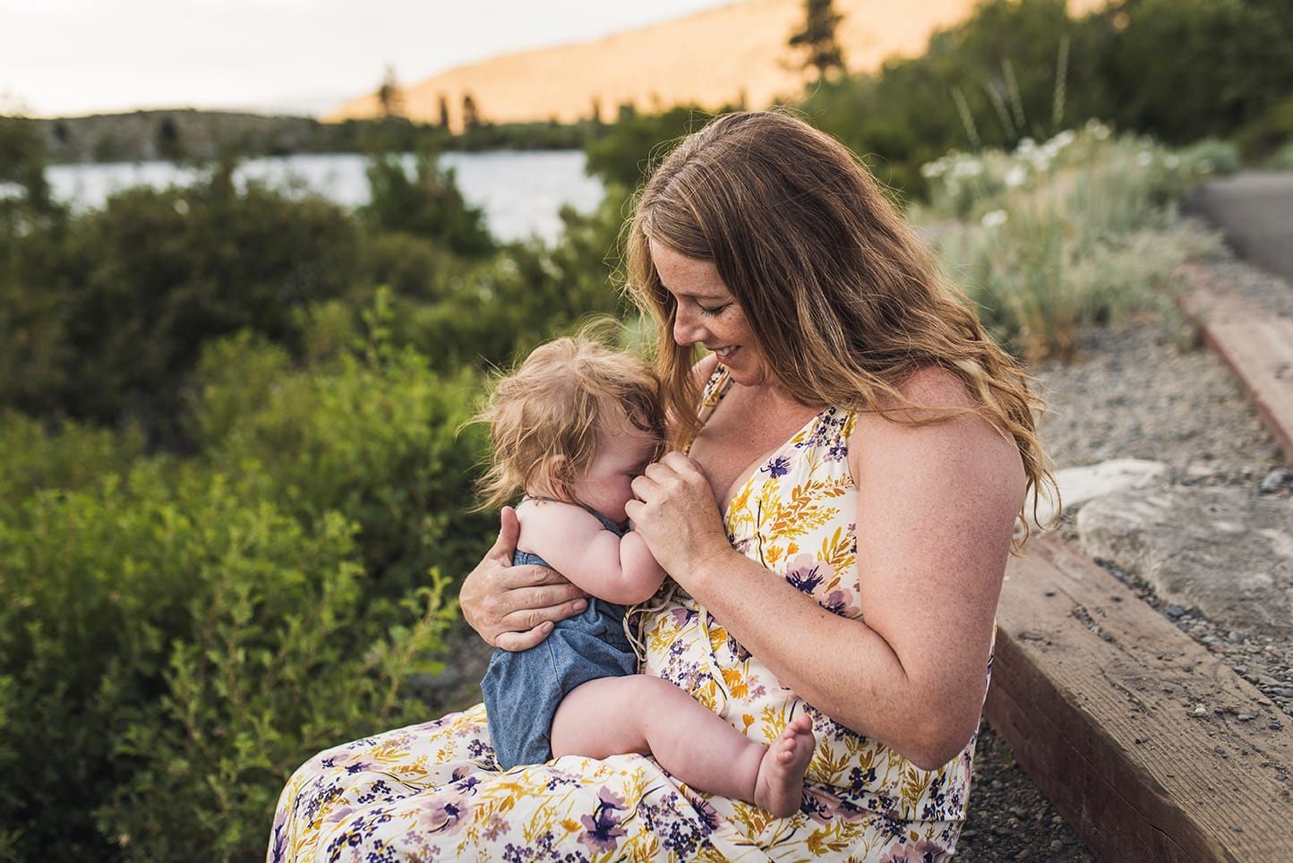 Mother sits in nature with a young child on her lap, breastfeeding