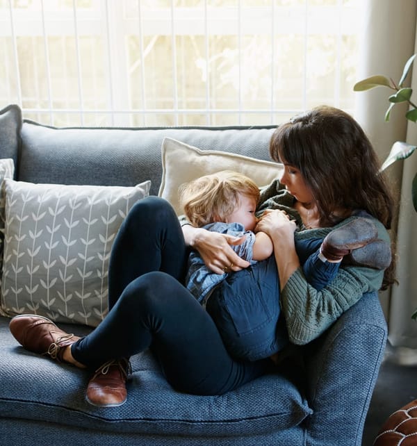 Photograph shows a mother breastfeeding her older child. They are curled up on a sofa