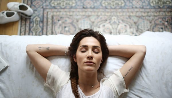 Woman with tattoos on her inner arms, reclines at the end of a bed