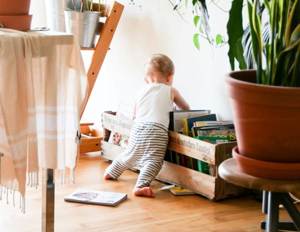 Photograph of a living space with plants and crates filled with books, with a toddler looking at the books