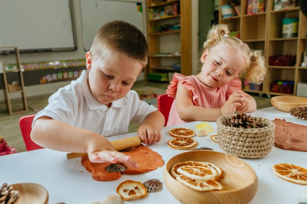 Two children sit at a table playing with coloured play dough and natural objects