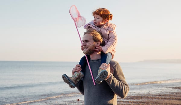 Photograph of a father carrying his daughter on his shoulders by the sea. She is holding a fishing net.