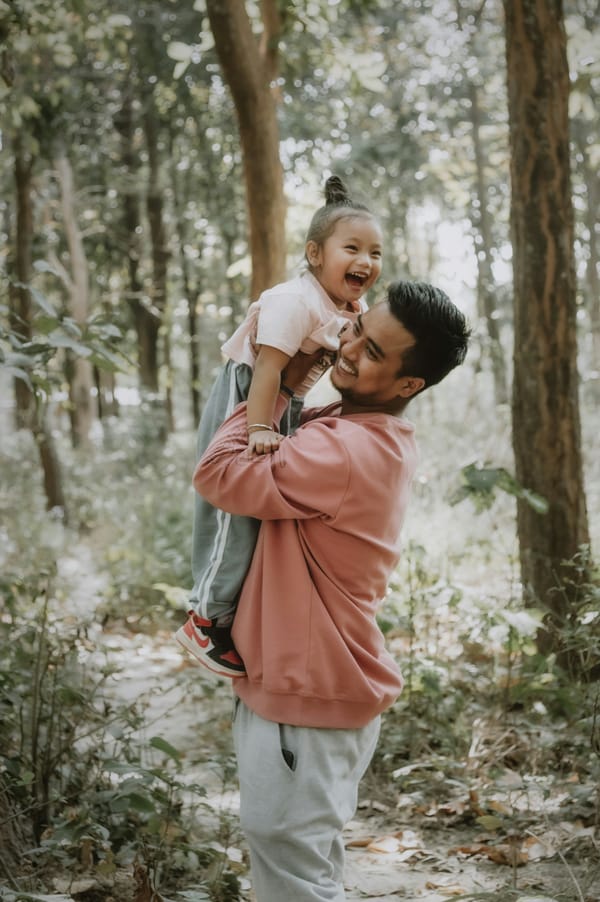 Photograph of a laughing child with her father; he is holding her up in the air, they're on a path through the trees